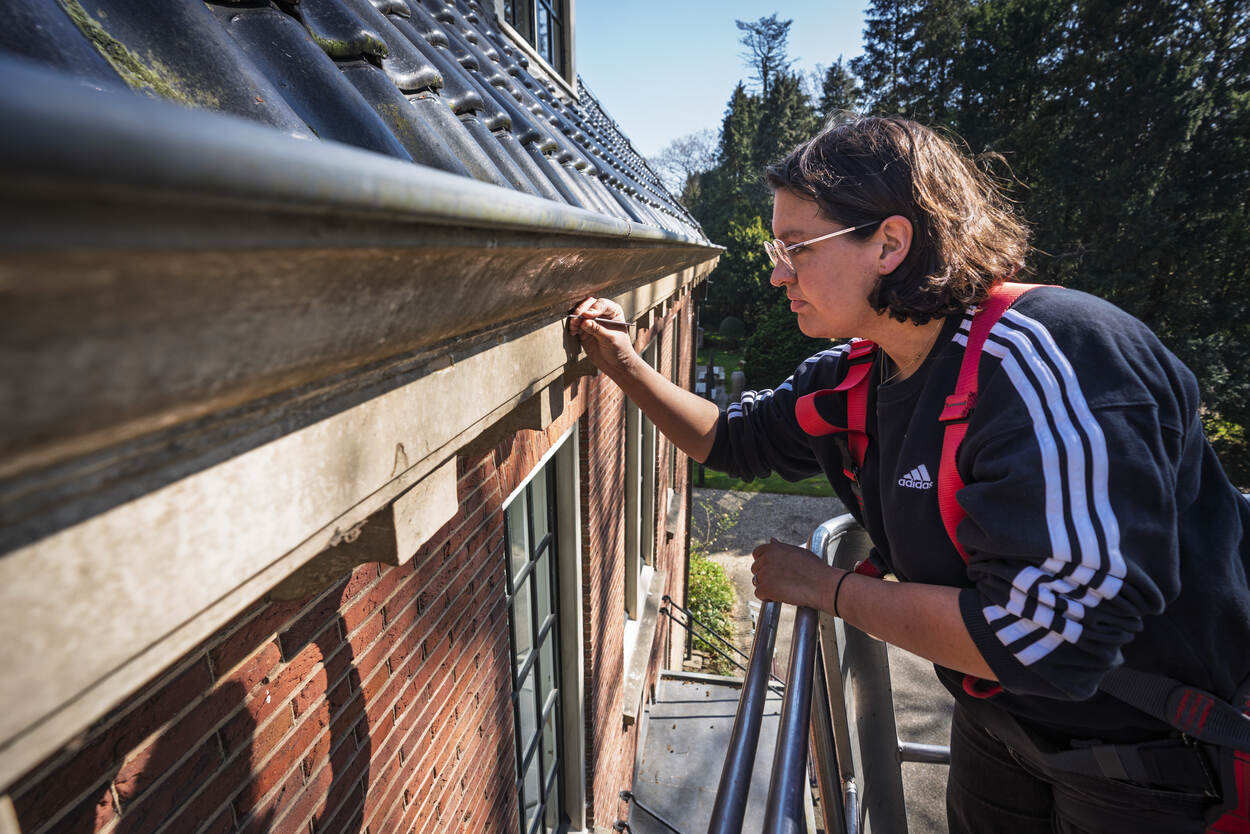 Lynn van Rijnsoever is aan de buitenzijde van de villa bezig met het kleurhistorisch onderzoek vanuit een hoogwerker.